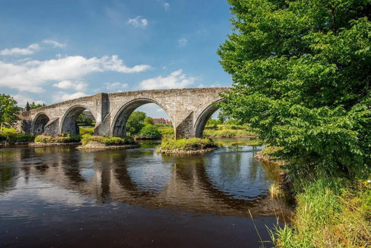 Old stone arch bridge over calm river surrounded by green trees under blue sky