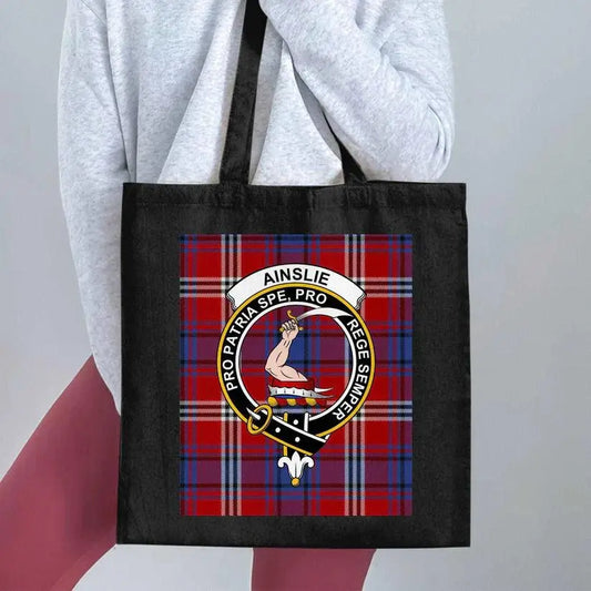 Black tote bag featuring Ainslie clan crest with red and blue tartan background, worn by person in gray top and pink pants