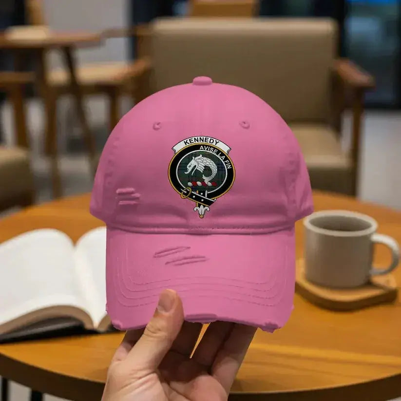 Hand holding pink distressed baseball cap with Kennedy clan crest patch on wooden table with book and mug