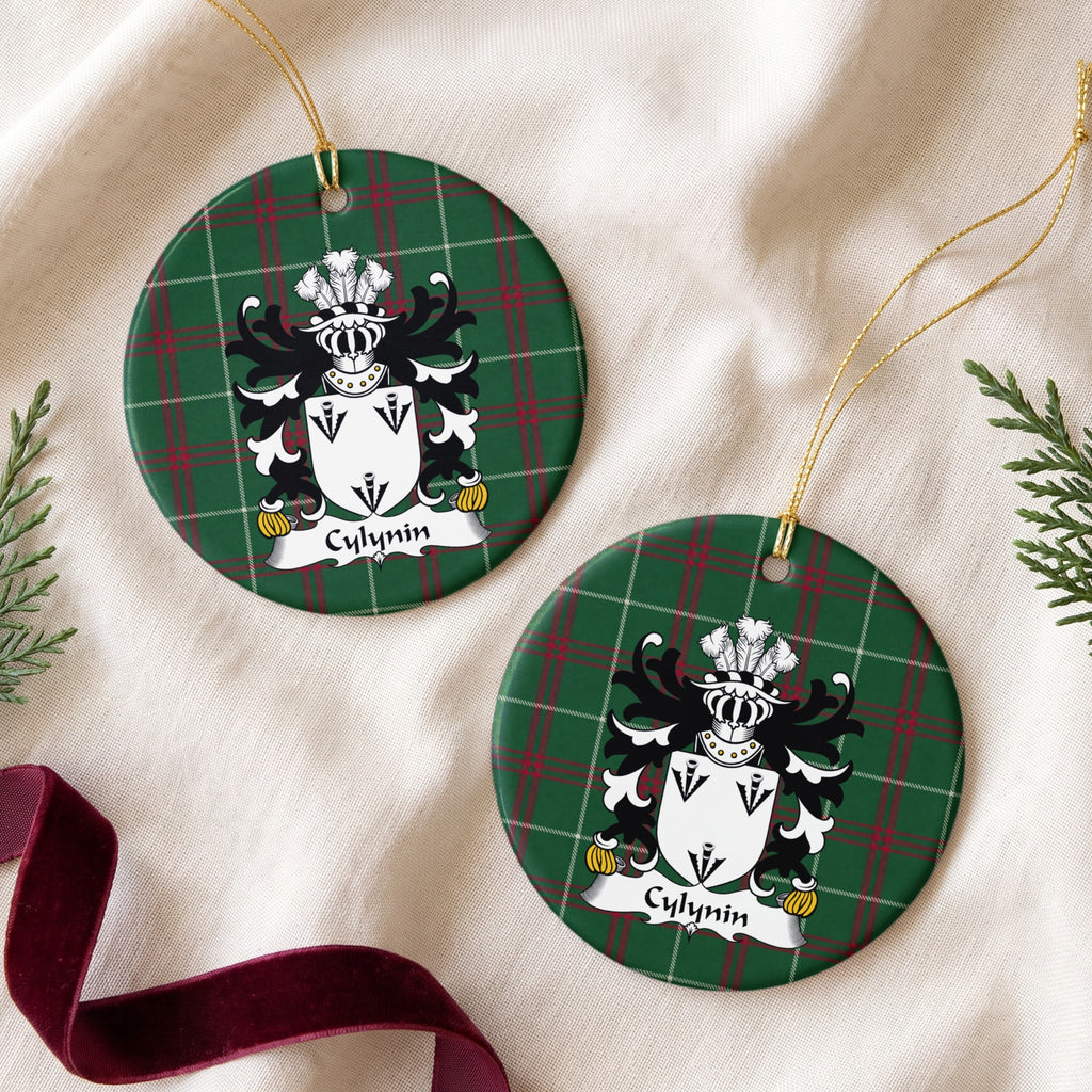 Two Cylynin ornaments on cream fabric beside evergreen and burgundy ribbon; three black arrowhead motifs on a white shield.