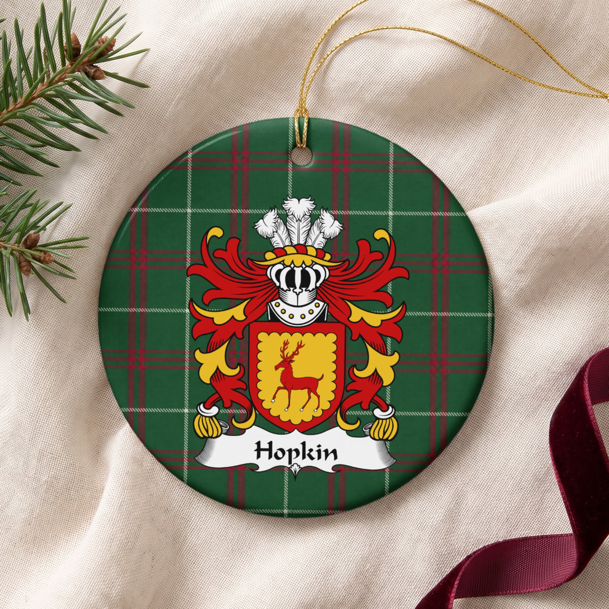 Hopkin ornament on cream fabric beside evergreen and burgundy ribbon, with gold string; a red stag on a gold shield with a scalloped red border.