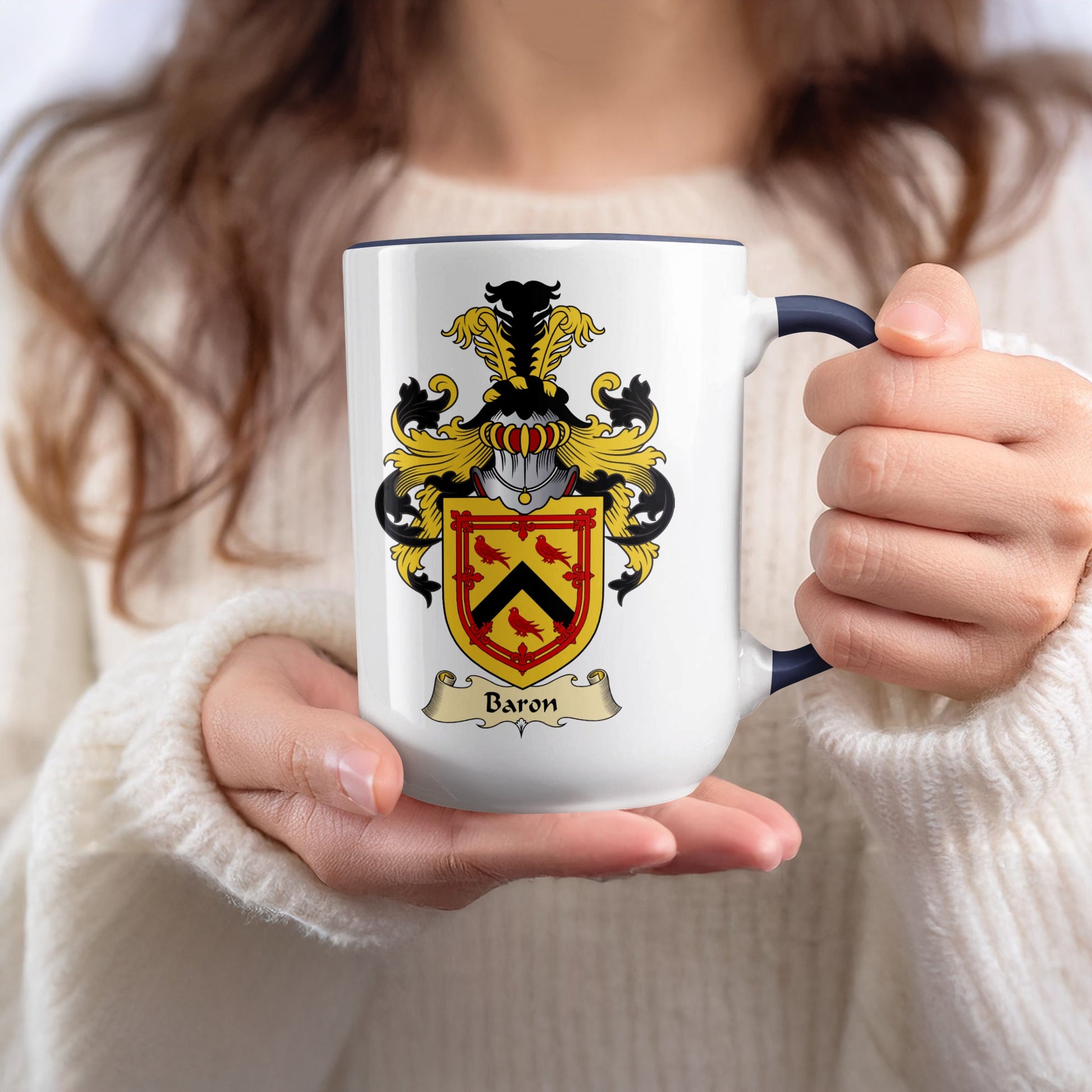 White mug with blue rim held by hands, featuring a family crest with a shield and a banner reading "Baron."