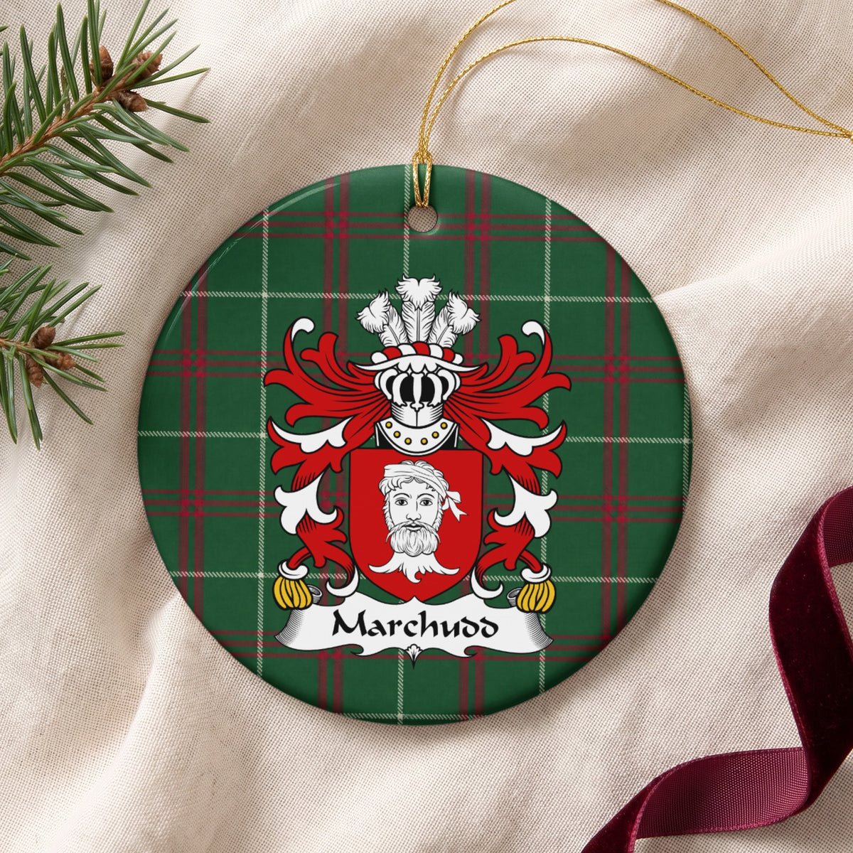 Marchudd ornament on cream fabric with gold hanging string, evergreen and burgundy ribbon; a red shield with a white bearded head on Welsh national tartan.
