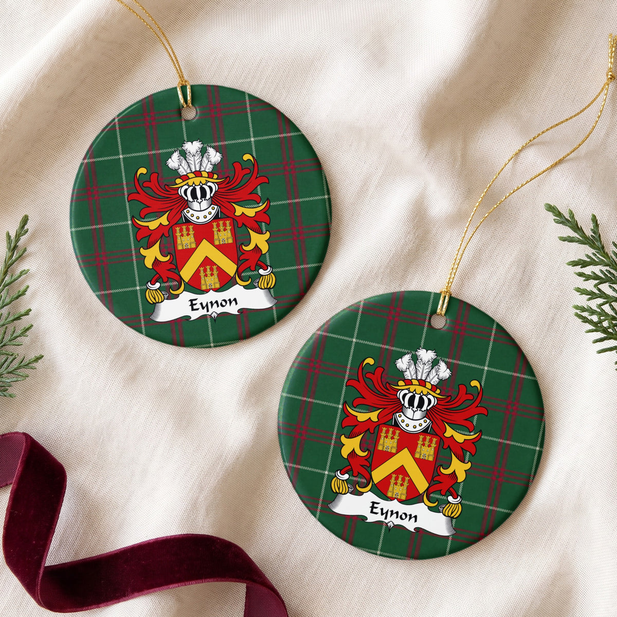 Two Eynon ornaments on cream fabric beside burgundy ribbon; a gold chevron between three gold castles on a red shield on Welsh national tartan.