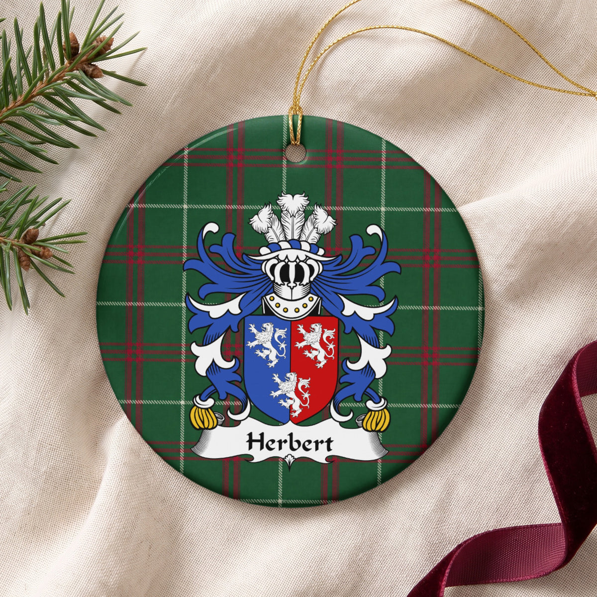 Herbert ornament on cream fabric beside evergreen and burgundy ribbon, with gold string; three white lions on a vertically divided blue-and-red shield.