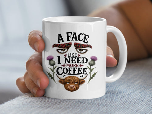 White mug with a grumpy Highland cow face and text "A face like I need more coffee."