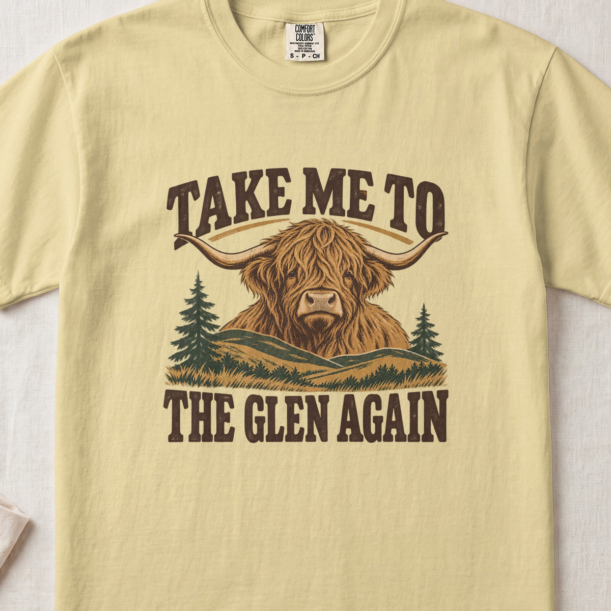 Take Me to the Glen Again Highland cow design on a Butter Comfort Colors 1717 T-shirt, flat lay