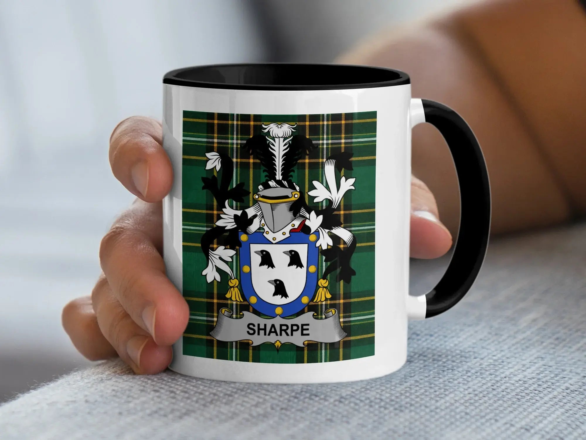 White mug with black rim held by a hand, displaying a green tartan shield with three birds.
