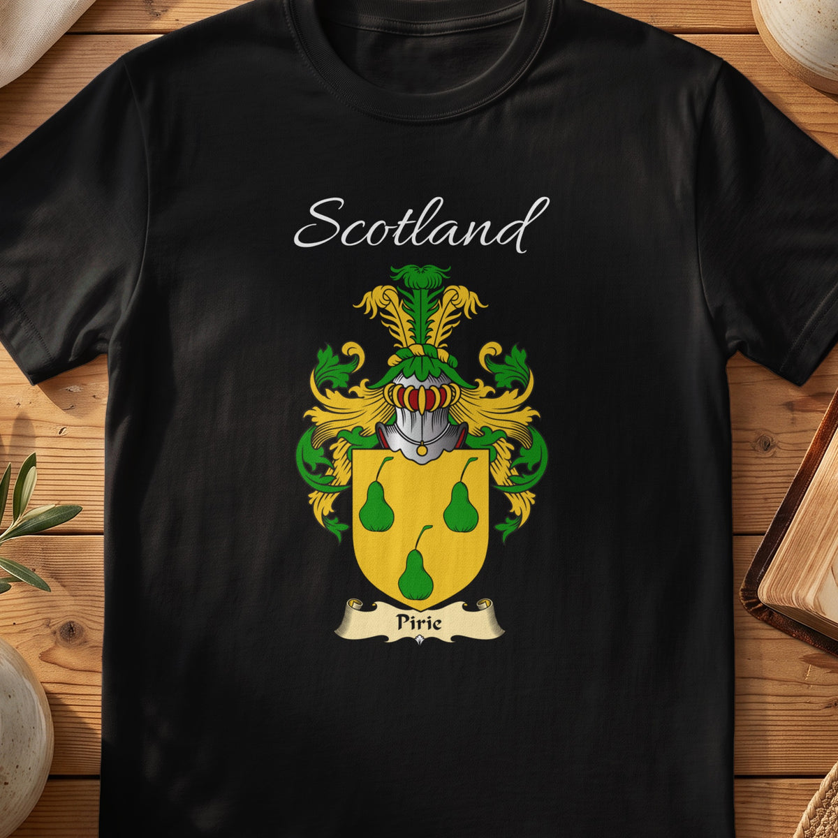 Black Pirie Scottish family crest T-shirt laid flat, showing “Scotland” above a green-and-gold family shield featuring three pear shapes.