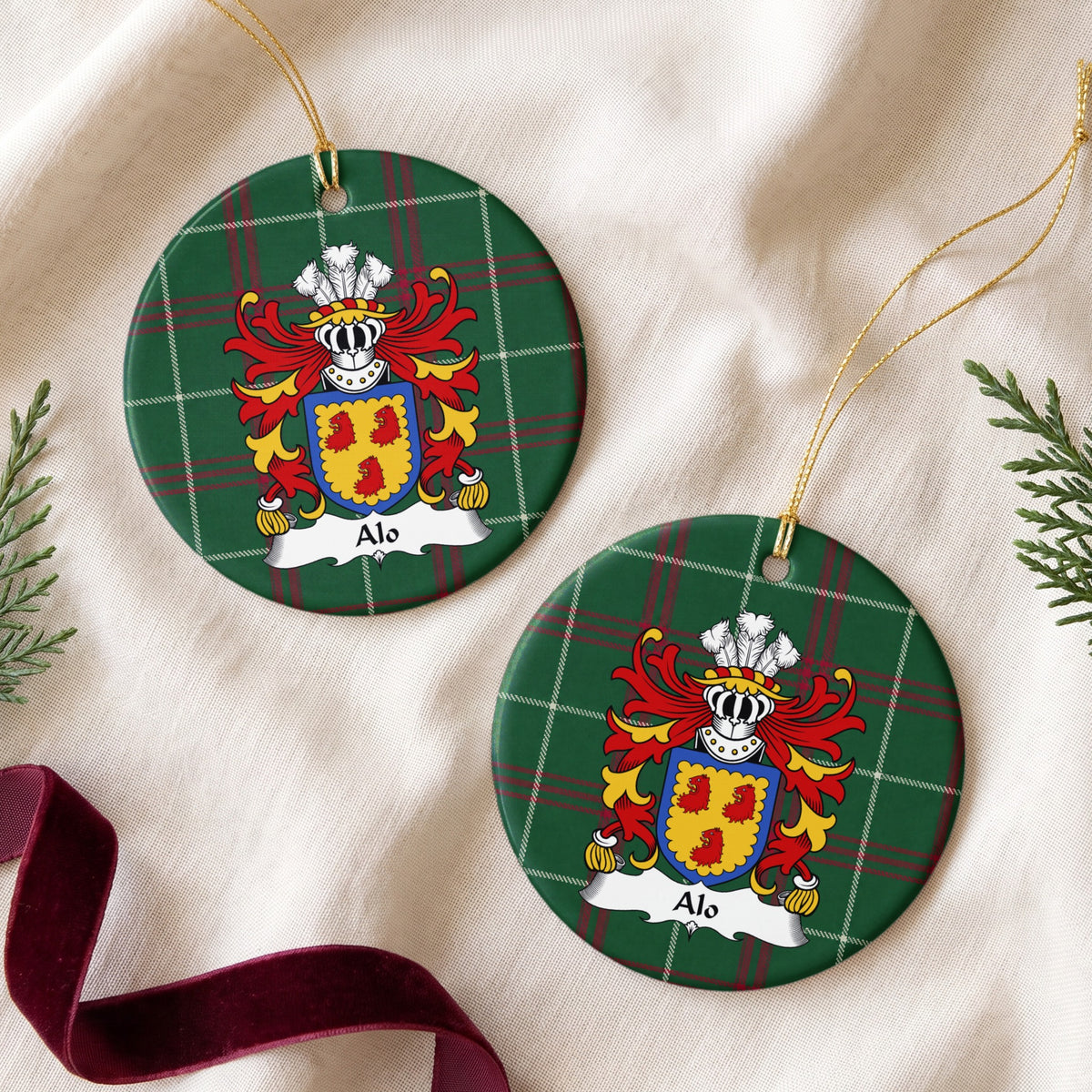 Two round ornaments showing the Alo family crest and surname on Welsh national tartan, arranged on cream fabric with gold cords, evergreen sprigs and burgundy ribbon.