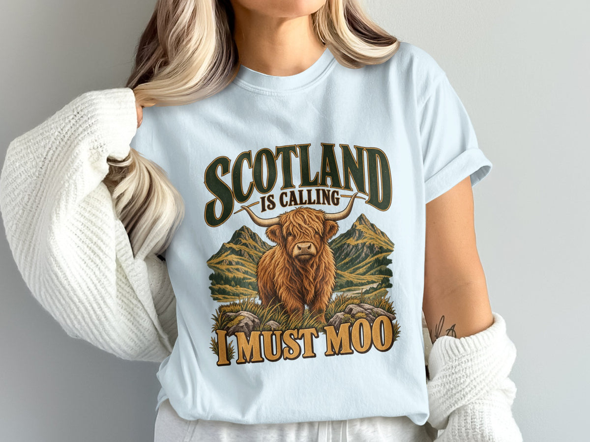 Model wearing the Scotland Is Calling, I Must Moo Highland cow Comfort Colors T-shirt in Chambray