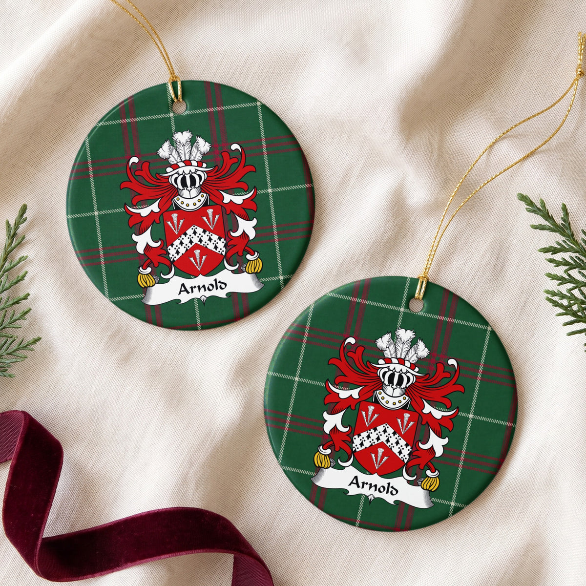 Two round ornaments showing the Arnold family crest and surname on Welsh national tartan, arranged on cream fabric with gold cords, evergreen sprigs and burgundy ribbon.