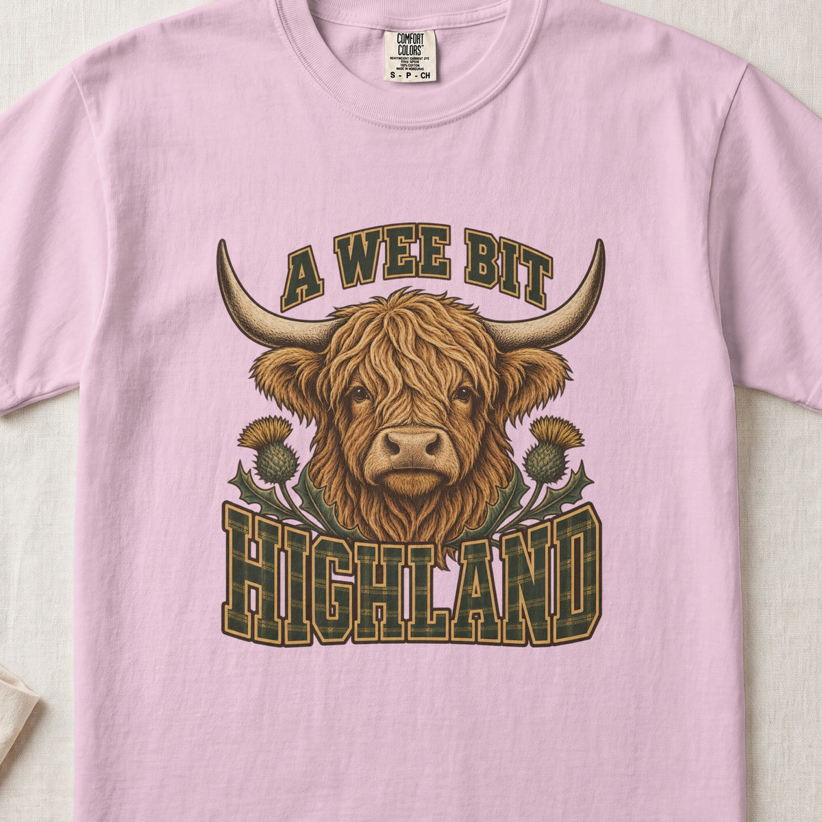 A Wee Bit Highland Highland cow design on a Blossom Comfort Colors 1717 T-shirt, flat lay