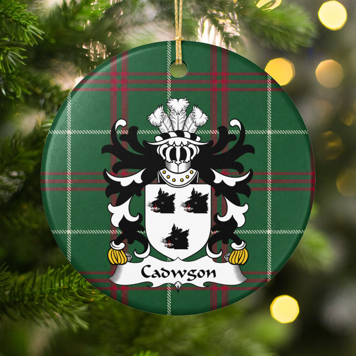 Cadwgon Style 1 ornament hanging among Christmas tree branches and warm lights; three black boar heads on a white shield on Welsh national tartan.