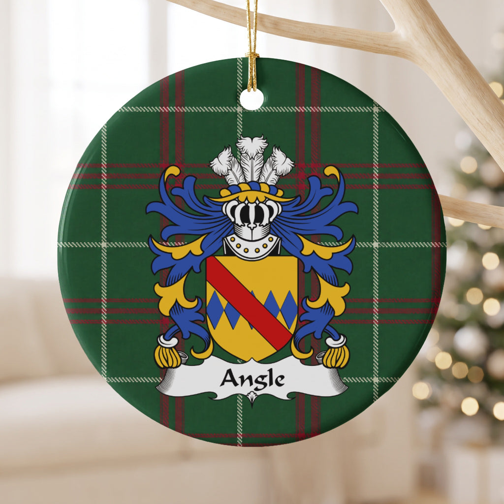 Angle family crest and surname on a round Welsh national tartan ornament, hanging by a gold cord from a pale wooden branch with blurred Christmas lights behind it.