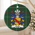 Angle family crest and surname on a round Welsh national tartan ornament, hanging by a gold cord from a pale wooden branch with blurred Christmas lights behind it.