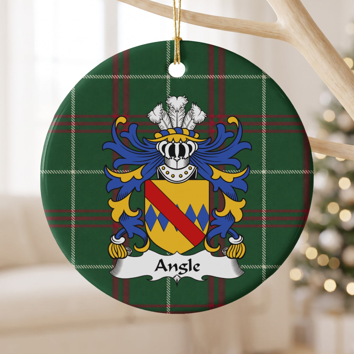 Angle family crest and surname on a round Welsh national tartan ornament, hanging by a gold cord from a pale wooden branch with blurred Christmas lights behind it.
