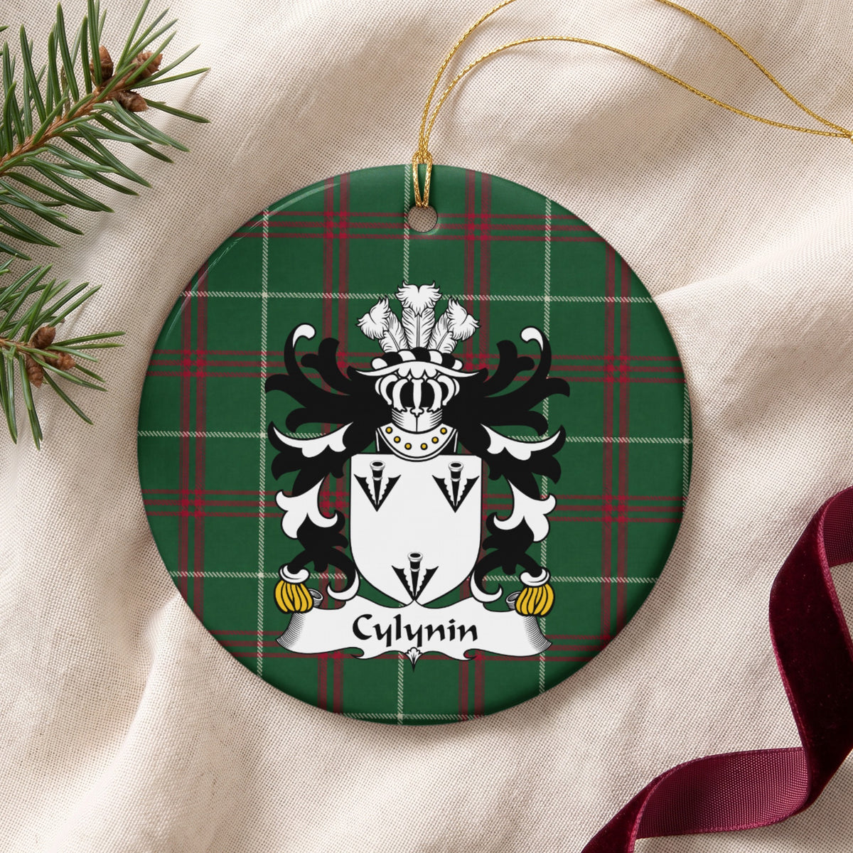Cylynin ornament on cream fabric with gold string, evergreen and burgundy ribbon; three black arrowhead motifs on a white shield.