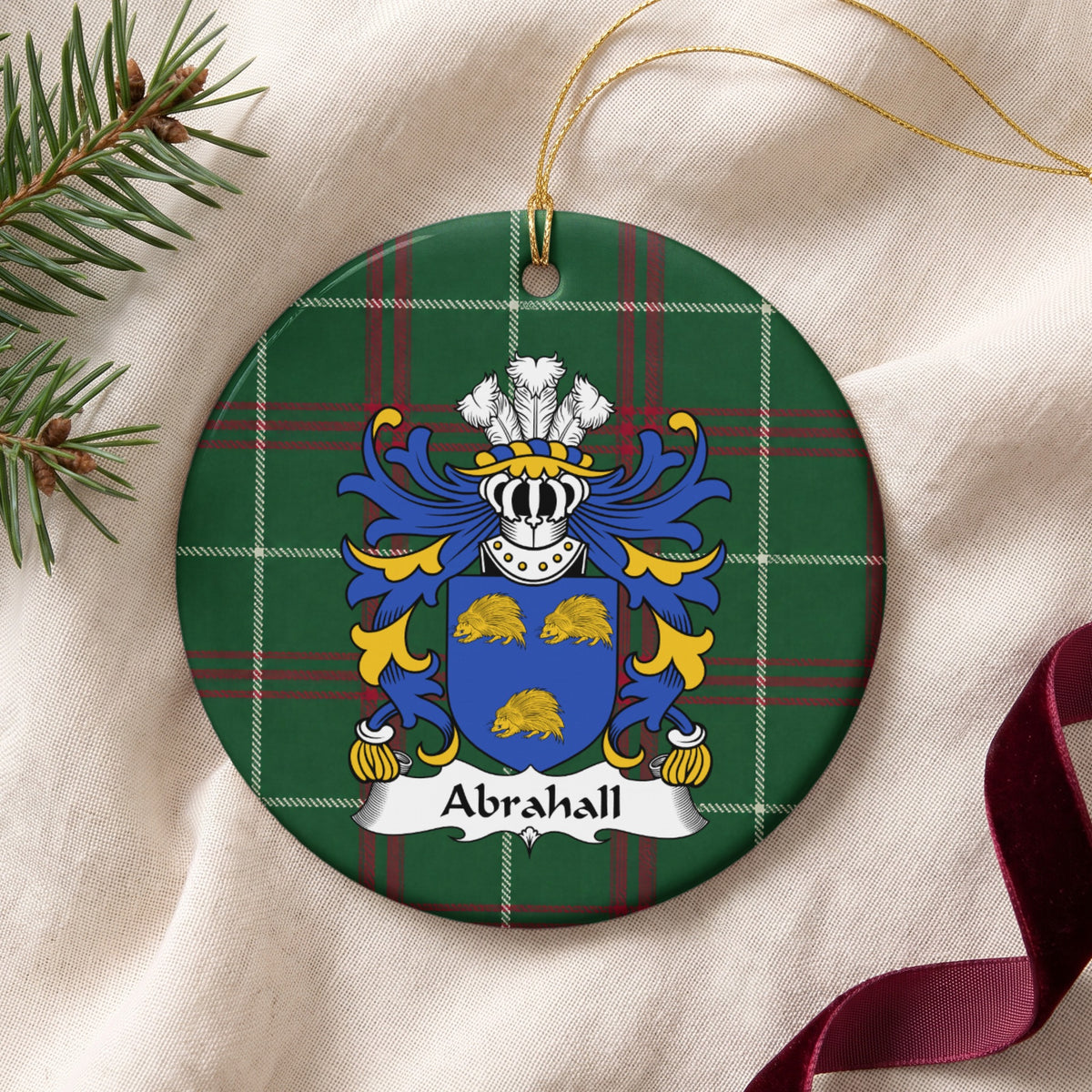 Abrahall family crest and surname on a round Welsh national tartan ornament with a gold cord, resting on cream fabric beside an evergreen sprig and burgundy ribbon.
