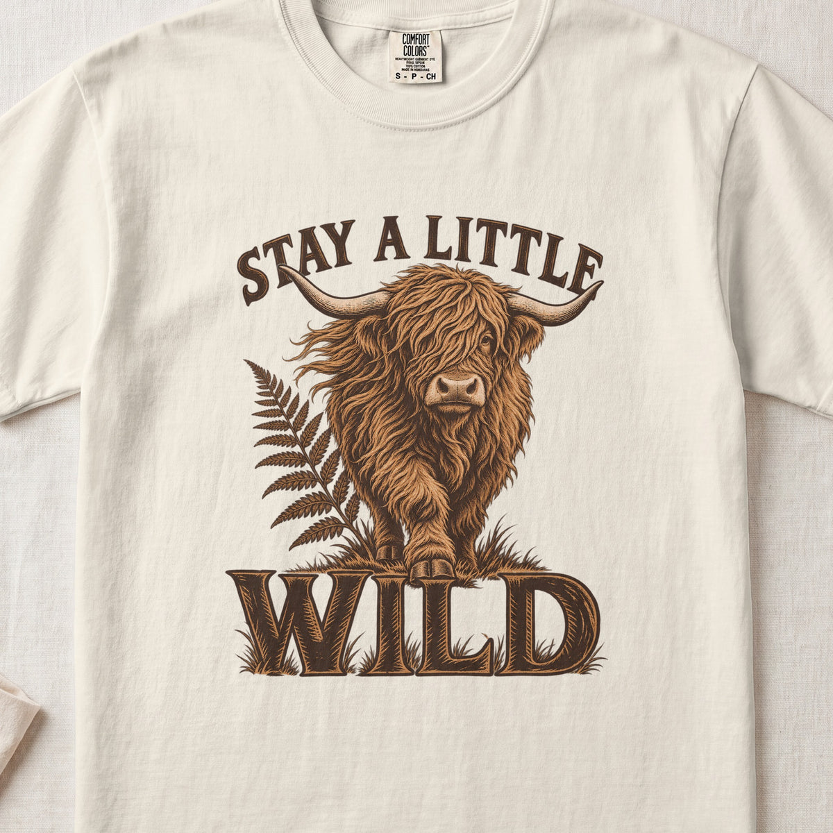 Stay a Little Wild Highland cow design on an Ivory Comfort Colors 1717 T-shirt, flat lay