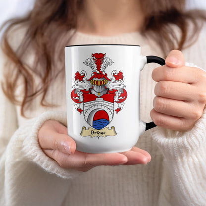 White mug with Bridge family crest held by person in cream sweater.
