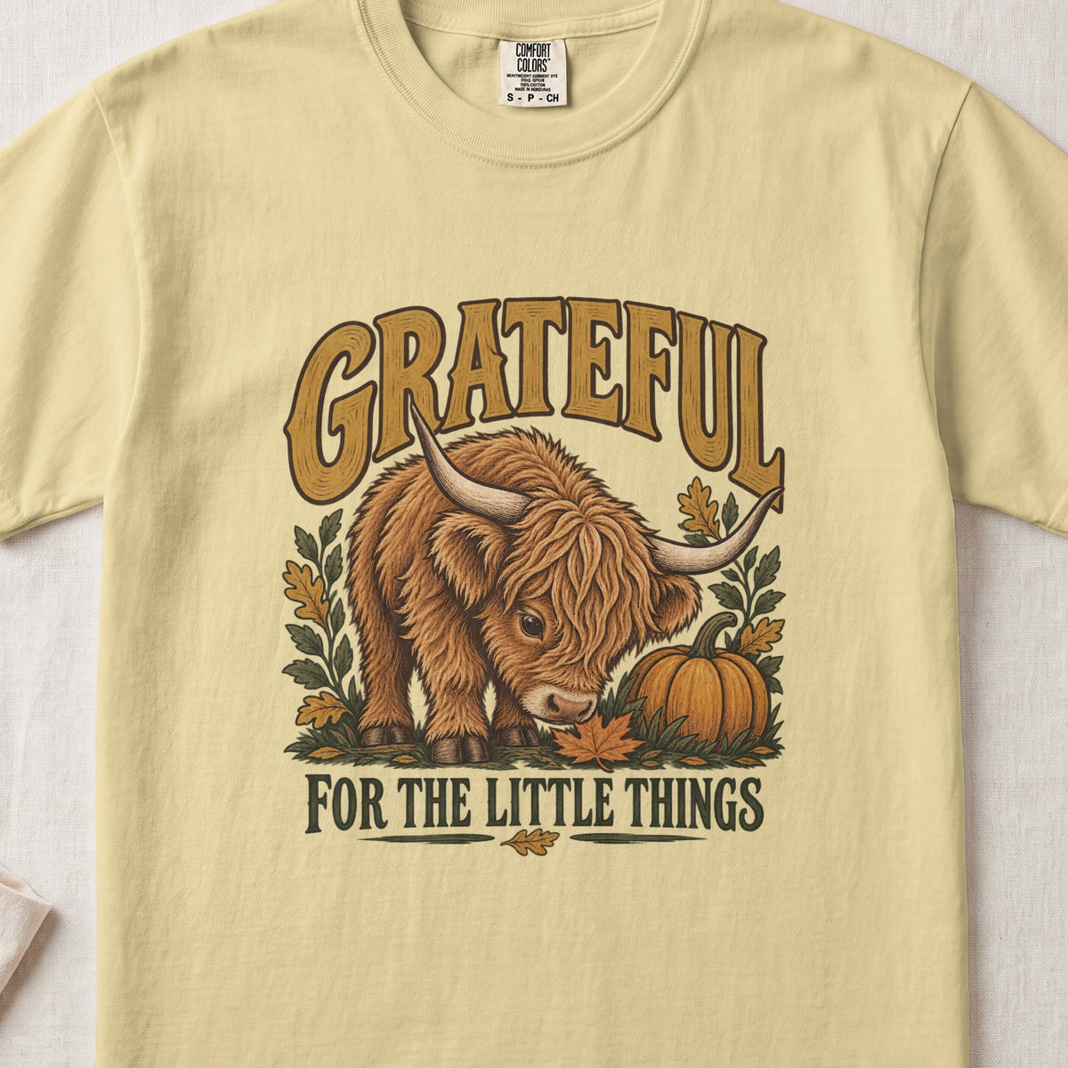 Grateful for the Little Things Highland cow design on a Butter Comfort Colors 1717 T-shirt, flat lay