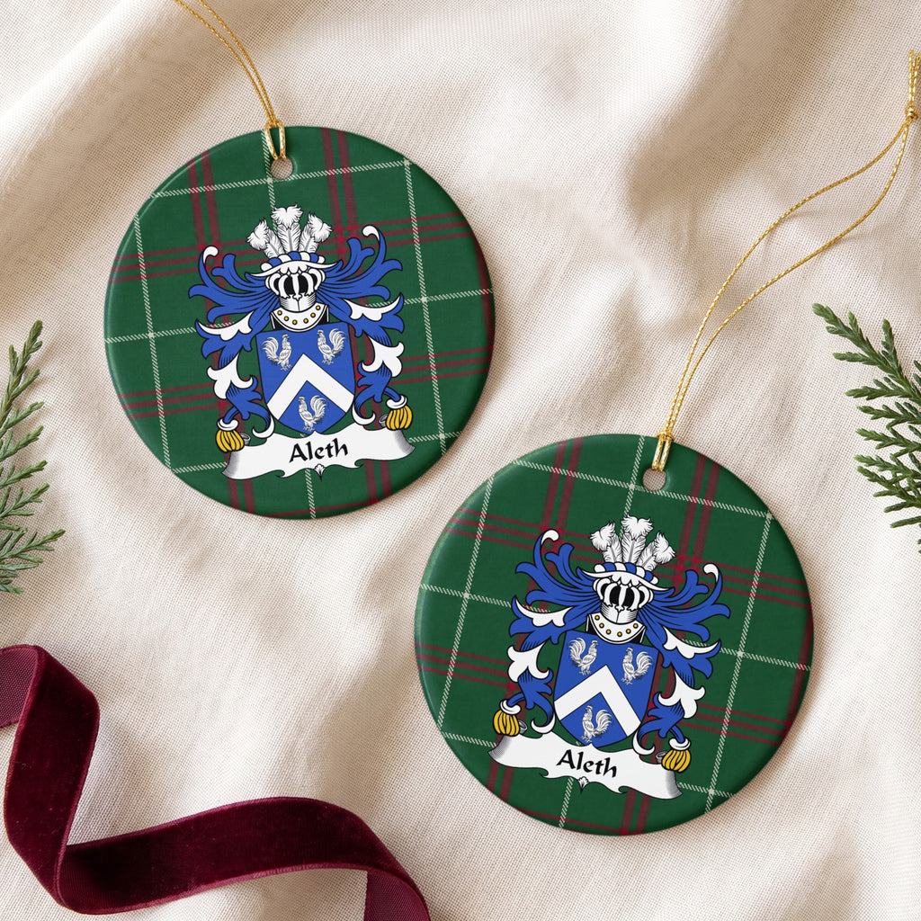 Two round ornaments showing the Aleth family crest and surname on Welsh national tartan, arranged on cream fabric with gold cords, evergreen sprigs and burgundy ribbon.