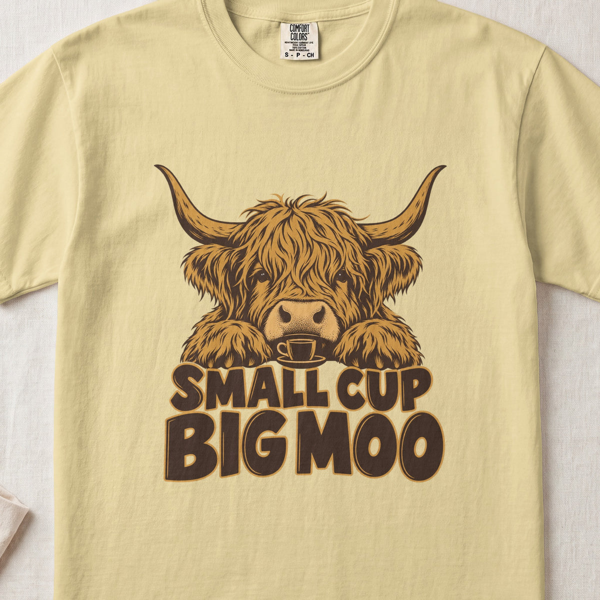 Small Cup, Big Moo Highland cow design on a Butter Comfort Colors 1717 T-shirt, flat lay