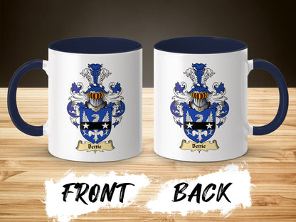 Two views of a white ceramic mug with a blue handle, featuring a coat of arms with the name "Bettie".