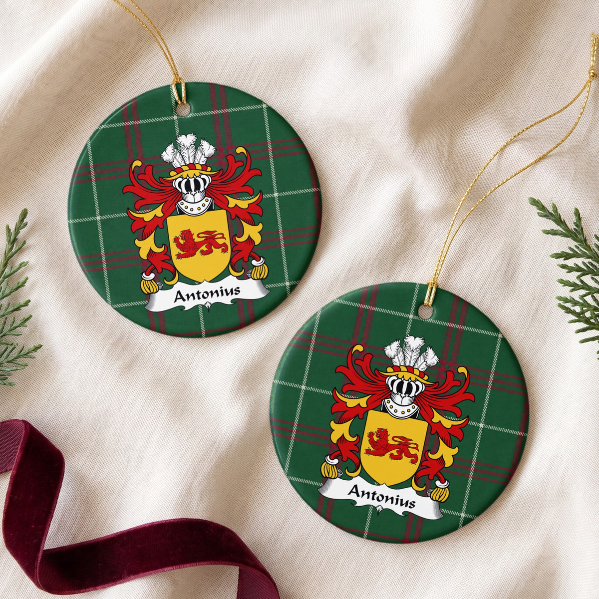 Two round ornaments showing the Antonius family crest and surname on Welsh national tartan, arranged on cream fabric with gold cords, evergreen sprigs and burgundy ribbon.