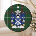 Aleth family crest and surname on a round Welsh national tartan ornament, hanging by a gold cord from a pale wooden branch with blurred Christmas lights behind it.