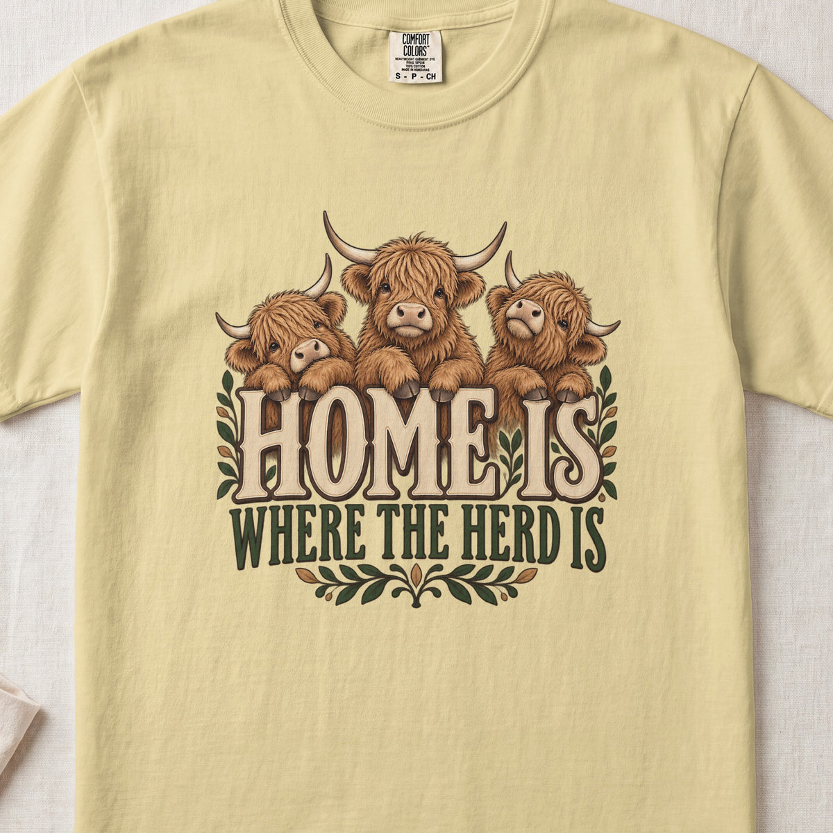 Home Is Where the Herd Is Highland cow design on a Butter Comfort Colors 1717 T-shirt, flat lay