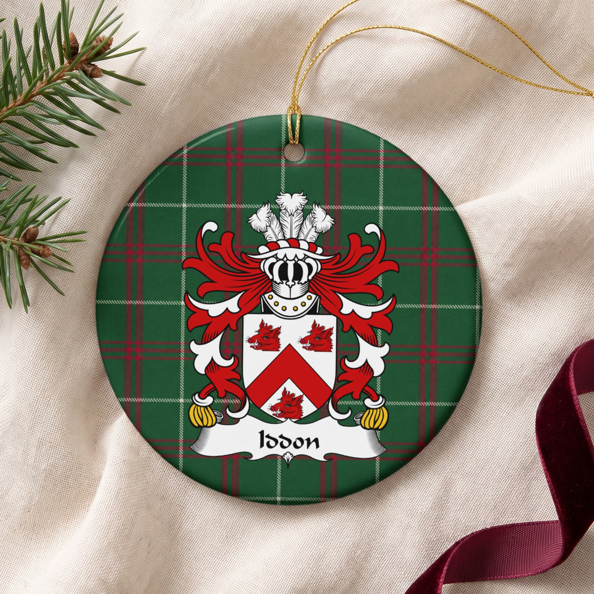 Iddon ornament on cream fabric with gold string, evergreen and burgundy ribbon; a red chevron and three red boar heads on a white shield on Welsh national tartan.