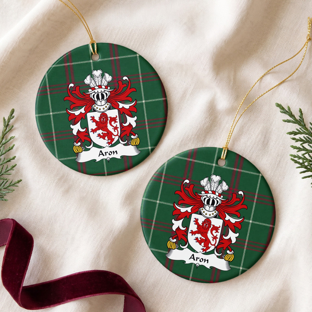 Two round ornaments showing the Aron family crest and surname on Welsh national tartan, arranged on cream fabric with gold cords, evergreen sprigs and burgundy ribbon.