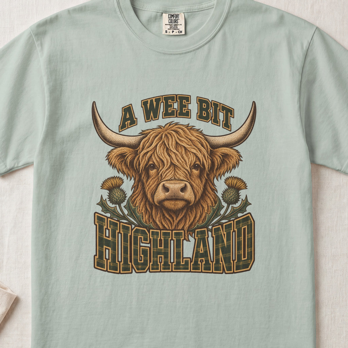A Wee Bit Highland Highland cow design on a Bay Comfort Colors 1717 T-shirt, flat lay