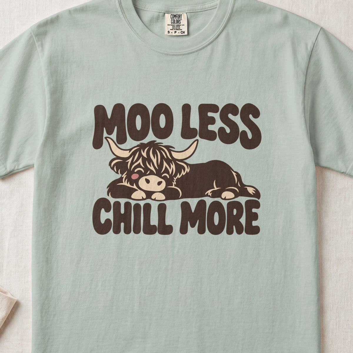 Moo Less, Chill More Highland cow design on a Bay Comfort Colors 1717 T-shirt, flat lay