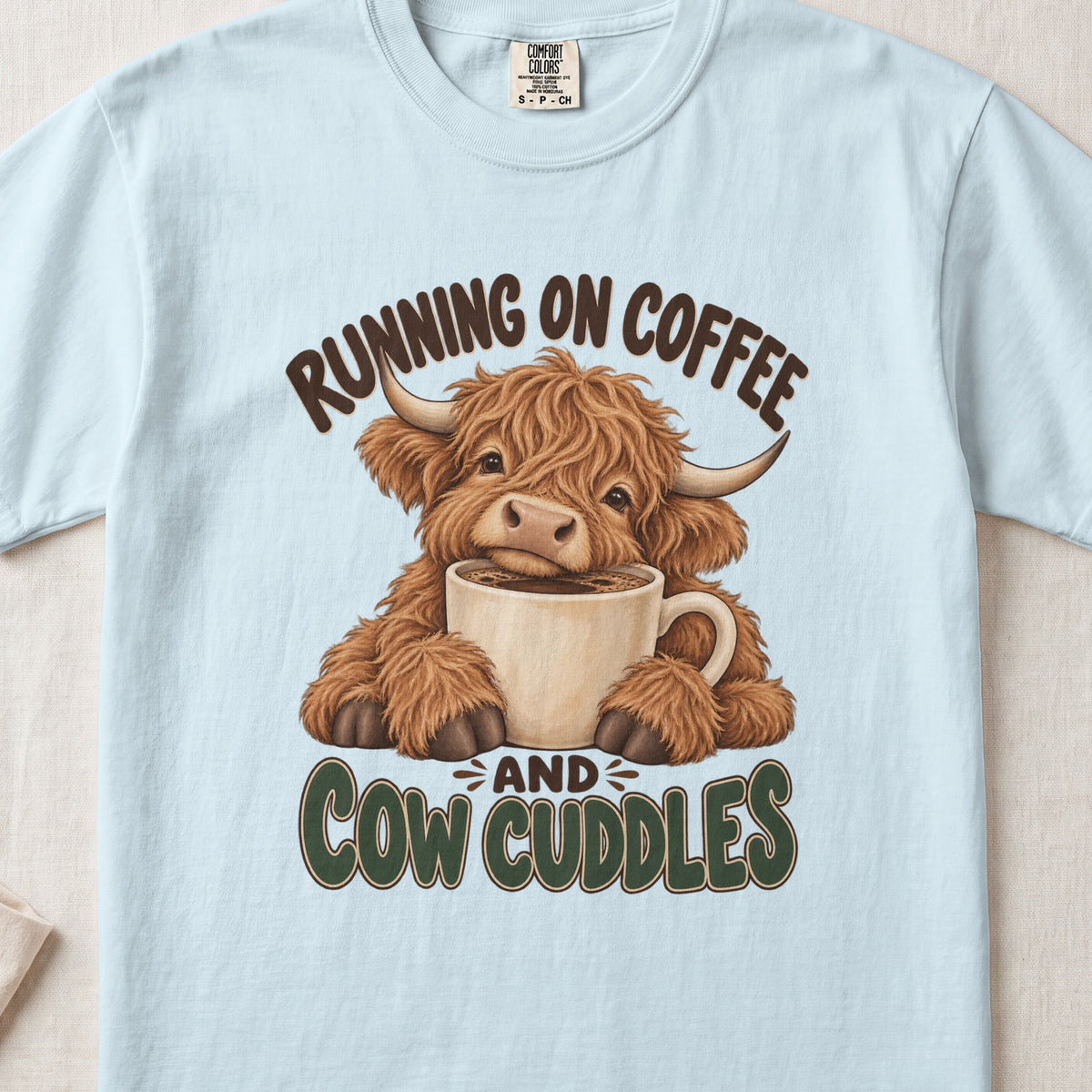 Running on Coffee & Cow Cuddles Highland cow design on a Chambray Comfort Colors 1717 T-shirt, flat lay