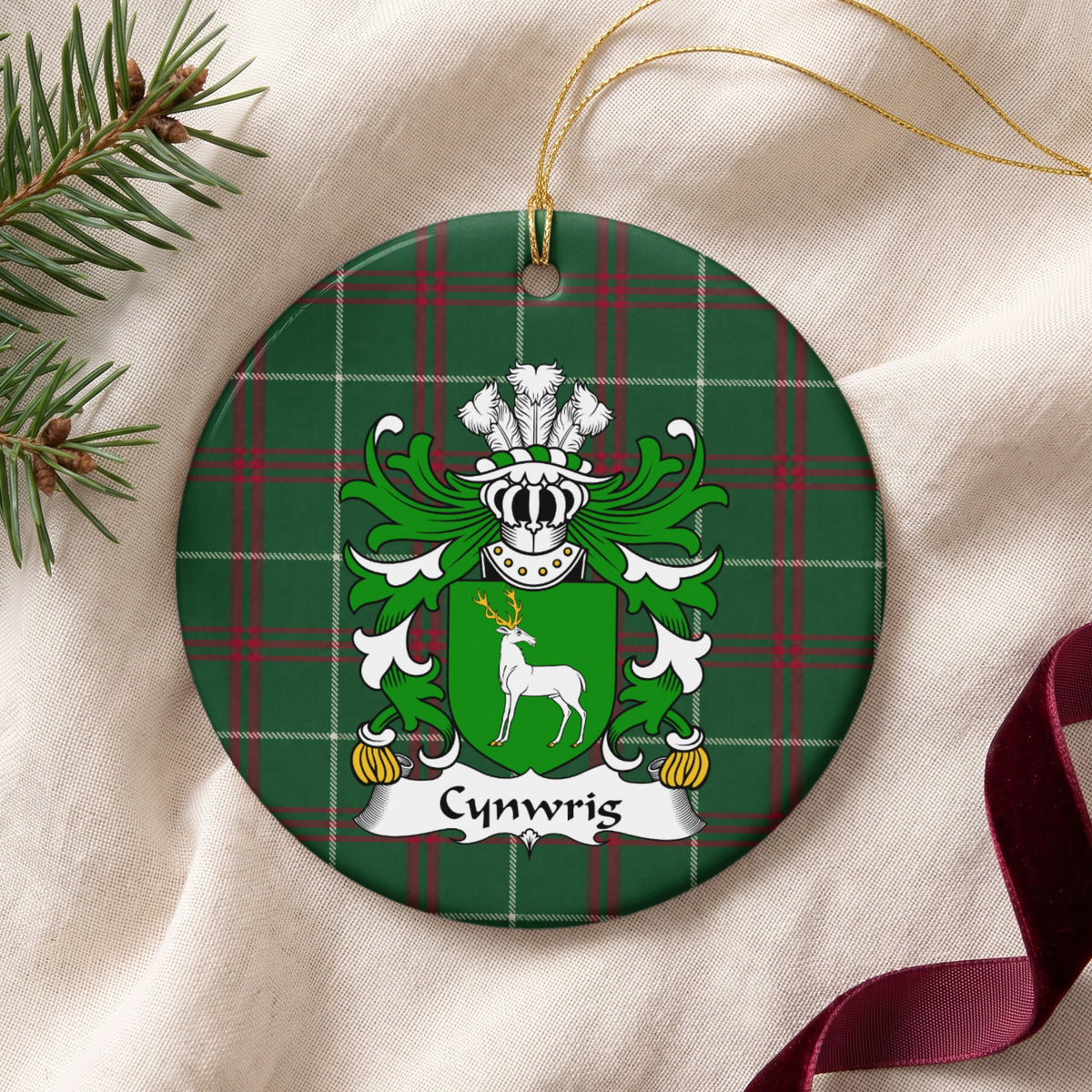 Cynwrig Style 2 ornament on cream fabric with gold string, evergreen and burgundy ribbon; a white stag with gold antlers on a green shield.