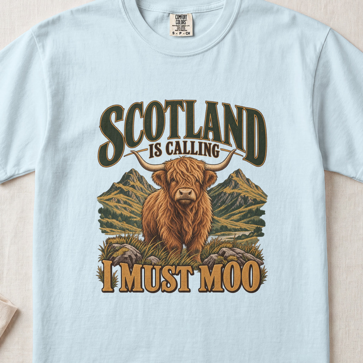 Scotland Is Calling, I Must Moo Highland cow design on a Chambray Comfort Colors 1717 T-shirt, flat lay