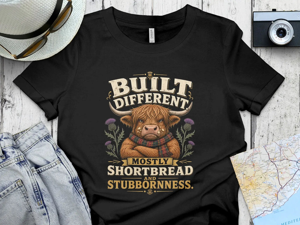 Black t-shirt with a Highland cow graphic and text "BUILT DIFFERENT MOSTLY SHORTBREAD AND STUBBORNNESS."