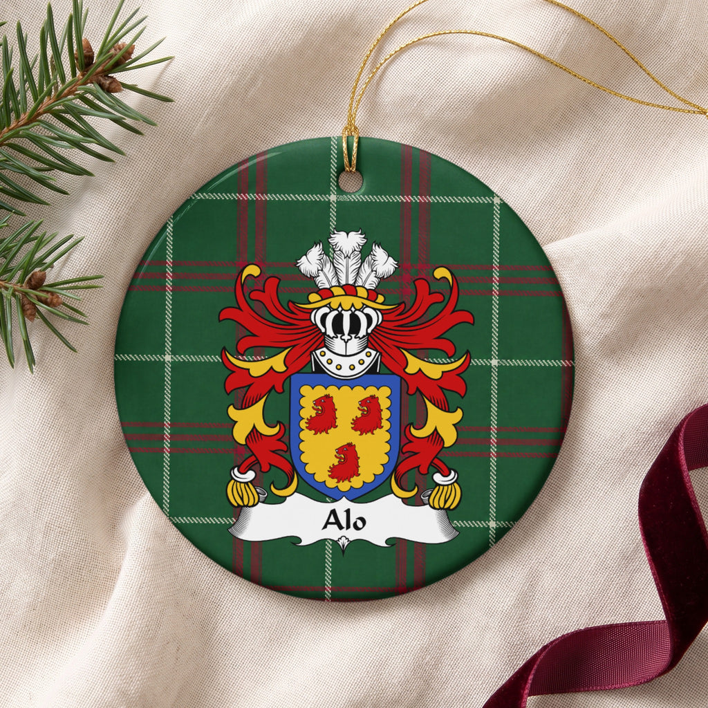 Alo family crest and surname on a round Welsh national tartan ornament with a gold cord, resting on cream fabric beside an evergreen sprig and burgundy ribbon.