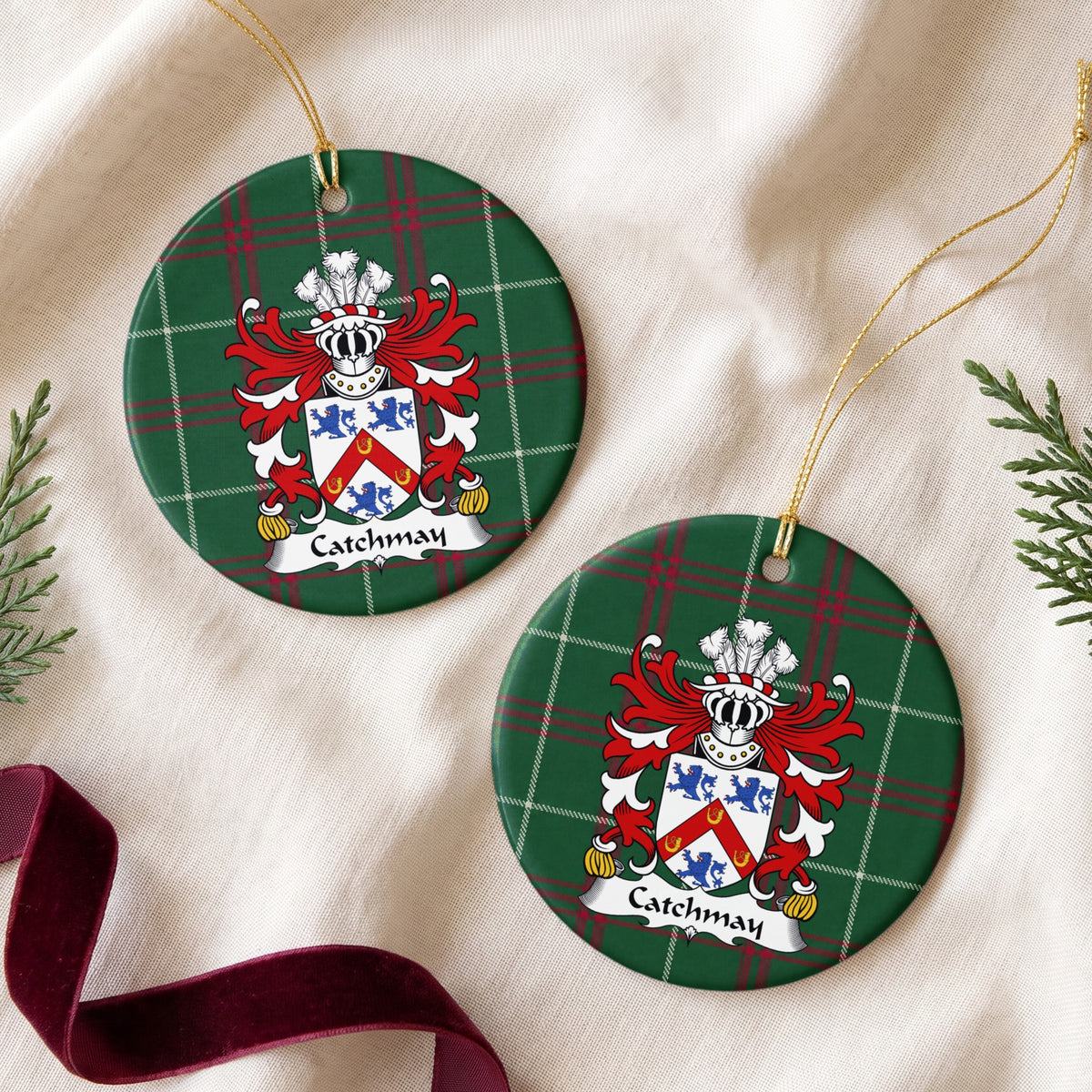 Two Catchmay ornaments on cream fabric beside evergreen sprigs and burgundy ribbon; a red chevron bearing three gold horseshoes between three blue lions on a white shield on Welsh national tartan.