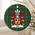 Alo family crest and surname on a round Welsh national tartan ornament, hanging by a gold cord from a pale wooden branch with blurred Christmas lights behind it.