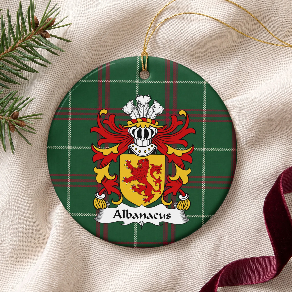 Albanacus family crest and surname on a round Welsh national tartan ornament with a gold cord, resting on cream fabric beside an evergreen sprig and burgundy ribbon.
