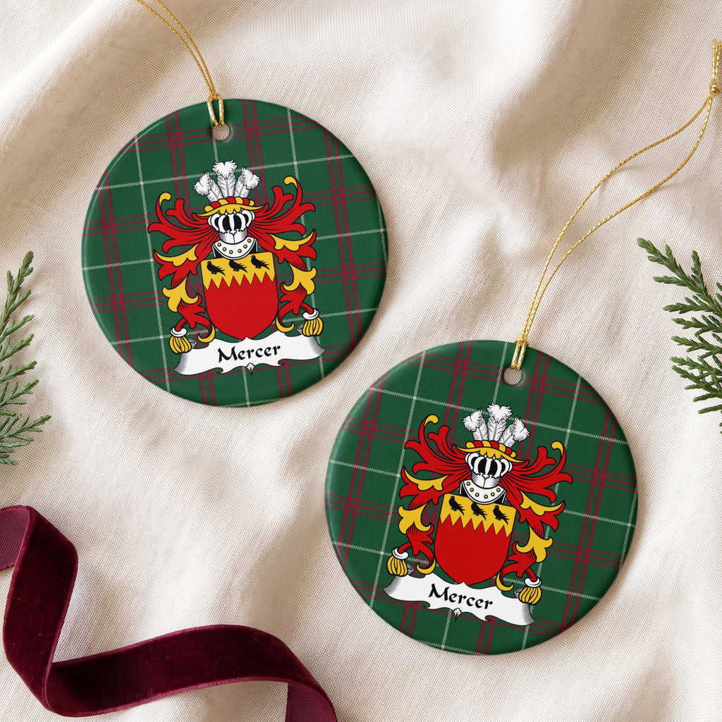Two Mercer ornaments on cream fabric beside evergreen and burgundy ribbon, showing three black birds on a jagged gold band above a red shield on Welsh national tartan.