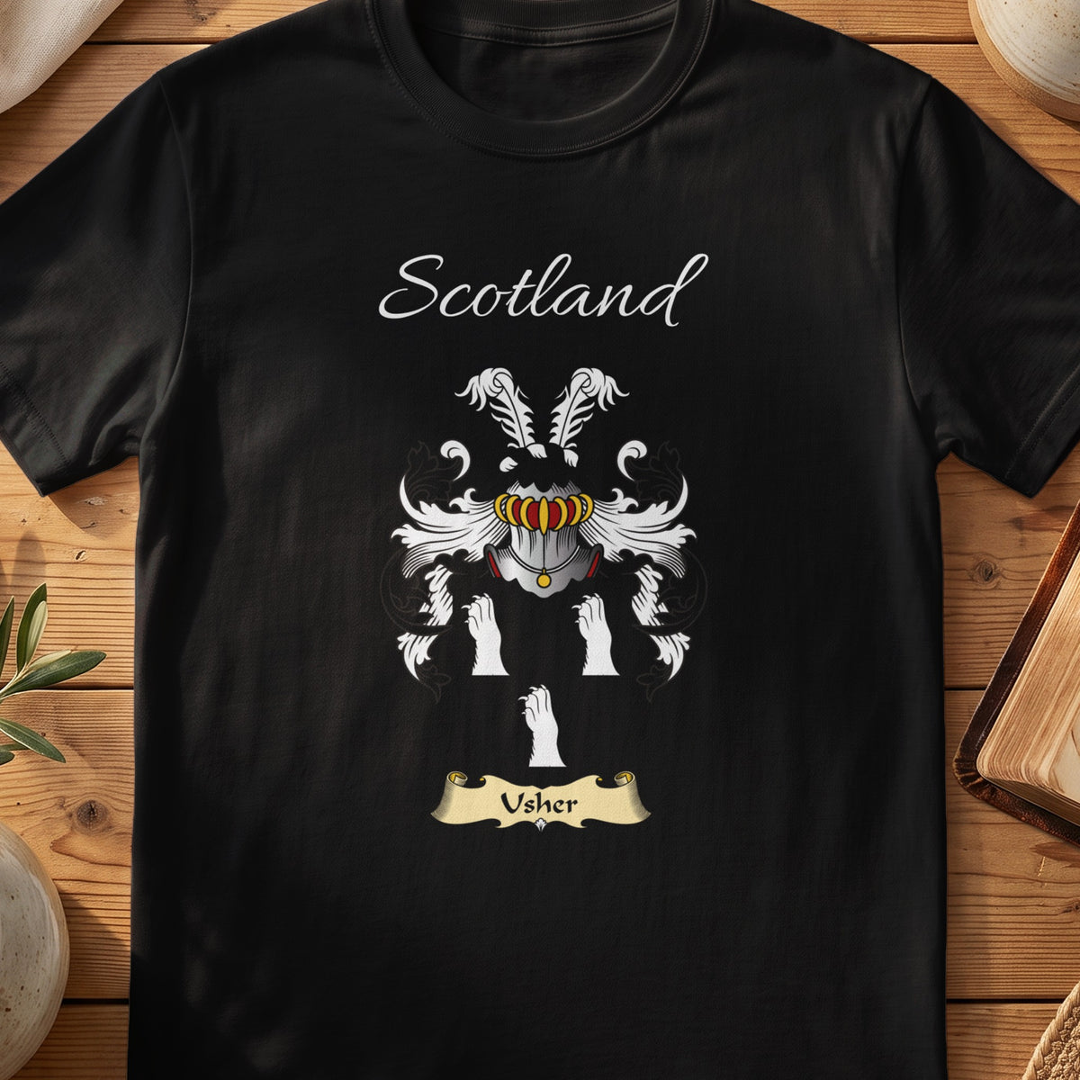 Black Usher Scottish family crest T-shirt laid flat, showing “Scotland” above a black-and-silver shield with paired goblet-like symbols.