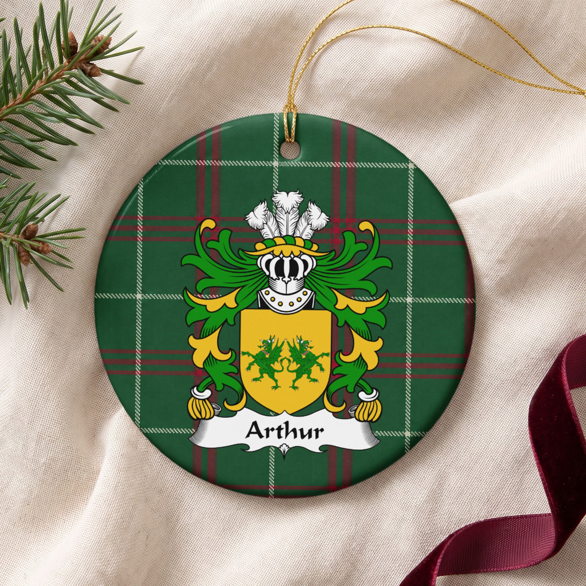 Arthur family crest and surname on a round Welsh national tartan ornament with a gold cord, resting on cream fabric beside an evergreen sprig and burgundy ribbon.