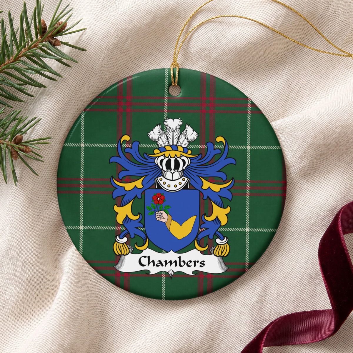 Chambers ornament on cream fabric with gold string, evergreen and burgundy ribbon; a hand in a gold sleeve holding a red rose on a blue shield on Welsh national tartan.