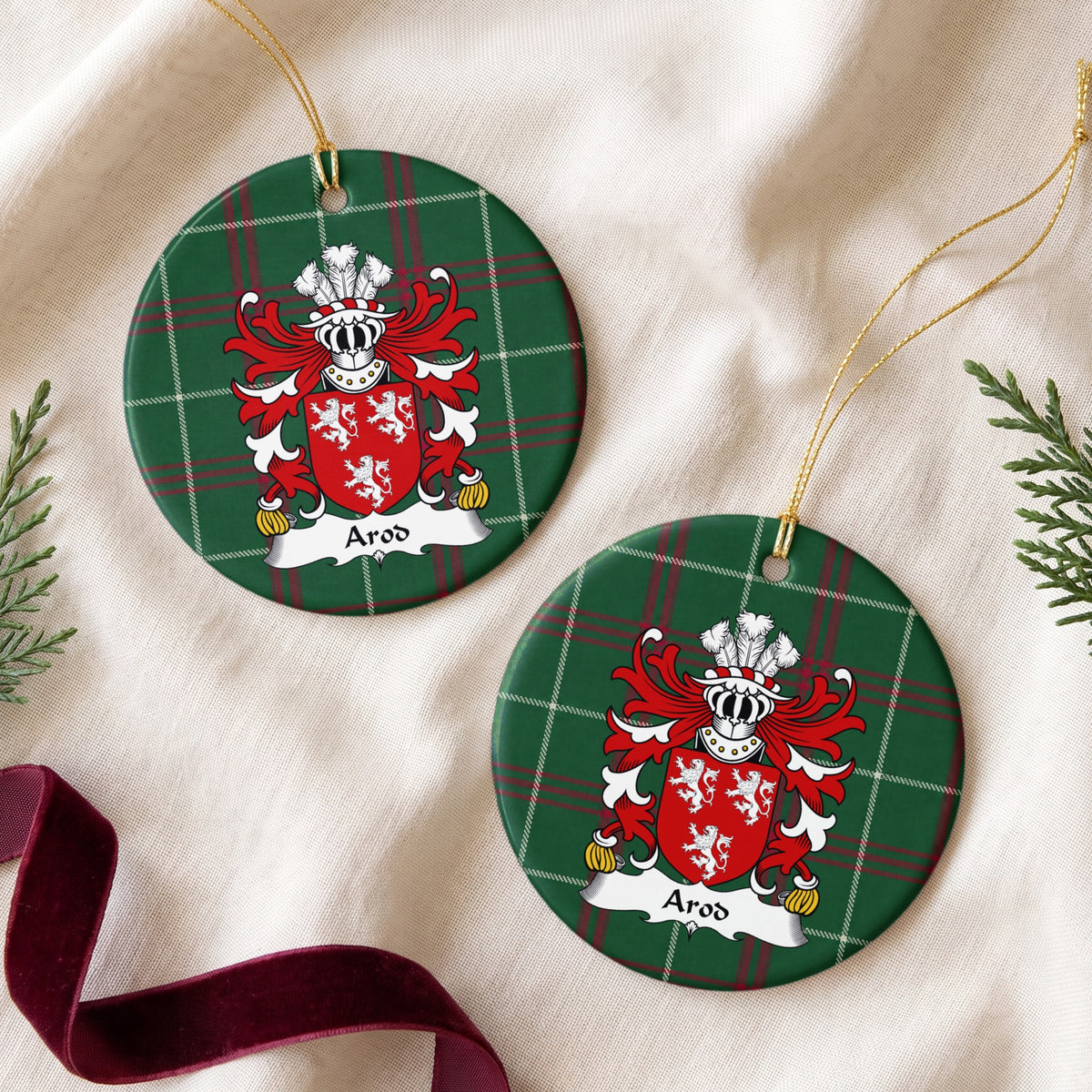 Two round ornaments showing the Arod family crest and surname on Welsh national tartan, arranged on cream fabric with gold cords, evergreen sprigs and burgundy ribbon.