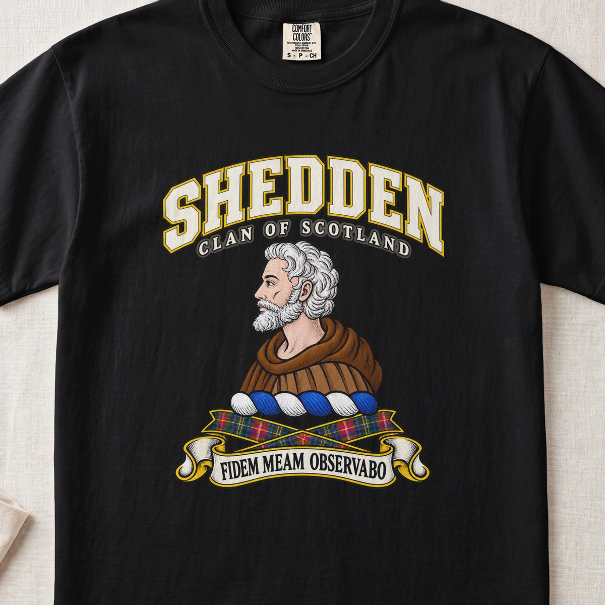 Shedden Scottish clan T-shirt in Black, front view with a gray-haired, bearded figure in a brown robe and motto design.